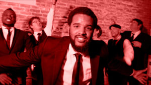 A man in a suit smiles at the camera while several other men in suits stand and cheer in the background, all under red lighting—an energetic scene that captures the spirit of a Boston wedding band performing by a brick wall.