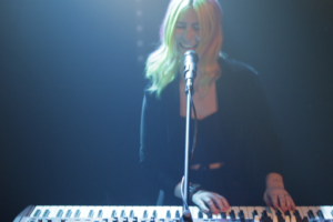 A person with light hair plays a keyboard and sings into a microphone on a dimly lit stage, performing as part of a Boston wedding band.