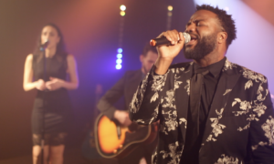 A man in a floral suit sings into a microphone on stage as part of a Boston wedding band, with a woman backup singer and a guitarist playing behind him under vibrant stage lights.
